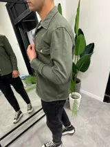 Sobrecamisa verde militar de corte recto, con cuello clásico, cierre frontal con botones metálicos, dos bolsillos con solapa en el pecho y mangas largas con puños abotonados.