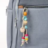 Mochila gris con detalles en marrón, con cierre superior y bolsillo frontal con cierre. Tiene manija corta superior y correas traseras regulables. Incluye llavero decorativo con cuentas de colores.