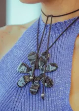 Collar con dije grande en forma de flor de seis pétalos con diseño abstracto en tonos grises y negros. El collar es de cordón negro con cuentas plateadas en los extremos.