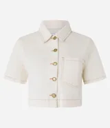 Camisa cropped de mangas cortas color crudo, confeccionada en sarga con cuello clásico, cierre frontal con botones dorados, bolsillo de parche en el pecho y costuras a tono.
