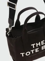 Bolso tote pequeño de lona color beige con estampado frontal en blanco con la leyenda "The Tote Bag Marc Jacobs". Tiene doble asa de mano y correa bandolera negra ajustable y extraíble.