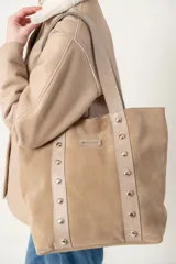 Bolso tipo tote confeccionado en cuero vacuno color beige, con tiras de cuero con tachas plateadas en el frente y logo de la marca grabado en el centro.