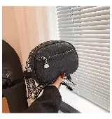 Cartera bandolera pequeña color beige, de cuero sintético con textura granulada. Tiene correa ancha de tela estampada con motivos geométricos y cierre superior con cremallera. Incluye un bolsillo frontal con cremallera y un dije metálico.