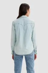 Camisa de denim celeste claro, de corte clásico, con cierre de botones a presión, mangas largas con puños abotonados, cuello americano y dos bolsillos frontales con solapa abotonada.