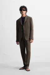 Pantalón de vestir color marrón oscuro, confeccionado en lino italiano. Presenta corte regular fit, cintura con pliegues, bolsillos laterales y traseros ribeteados, y terminación con dobladillo. Cierre frontal con cremallera y botón.