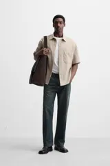 Sobrecamisa color beige de tejido elástico, con cuello solapa, manga corta y bolsillos de parche en el pecho. Cierre frontal con botones a presión.