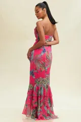 Vestido largo de tirantes finos, con escote drapeado y estampado de hojas tropicales en tonos verdes y azules sobre fondo fucsia. Tiene corte al bies y volantes en el ruedo.