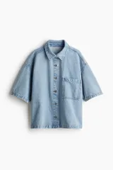 Camisa celeste de denim, corte amplio, con cuello clásico, manga corta, cierre frontal con botones metálicos, bolsillo de parche en el pecho y canesú en la espalda.