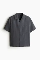 Camisa de manga corta color gris oscuro, confeccionada en mezcla de lino y algodón. Tiene cuello abierto, tapeta lisa, bolsillo de parche en el pecho y bajo recto con aberturas laterales.