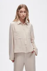 Camisa color natural de poliéster con cuello camisero, cierre con botones, dos bolsillos frontales y corte ancho.