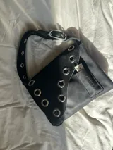 Cartera bandolera de cuero negro con ojalillos metálicos plateados, correa regulable con hebilla, bolsillo interior para labial y gancho para llaves. Diseño uruguayo.