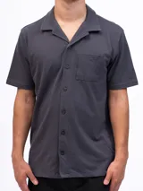 Camisa de manga corta confeccionada en tejido de piqué de algodón, color gris oscuro. Presenta un diseño de corte clásico con cuello tipo solapa, cierre frontal de botones al tono y un bolsillo de parche en el pecho.