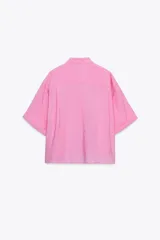 Camisa rosa de cuello solapa y manga corta con vuelta. Bajo asimétrico con aberturas laterales. Cierre frontal con botones ocultos por solapa.