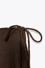 Pantalón marrón de tiro medio con cintura elástica y pierna ancha. Presenta una superposición tipo pareo ajustable con lazadas.