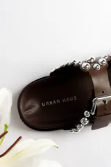 Sandalias marrones de cuero con dos tiras con hebillas ajustables y detalle de tachas plateadas.