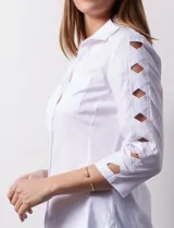 Camisa blanca de algodón con cuello clásico, solapa y escote en V. Tiene mangas tres cuartos con detalle de aberturas cruzadas. Presenta dos bolsillos delanteros aplicados y laterales en tejido modal.