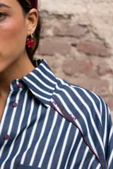 Camisa de rayas verticales azules y blancas, con cuello camisero, manga larga y puños abotonados. Presenta un bolsillo en el pecho con ribete rojo y un detalle de ribete rojo en el cuello y la tapeta de botones.