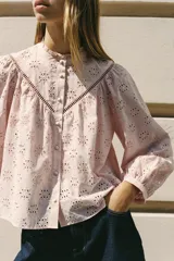 Camisa rosa pálido con bordados perforados, cuello con volante y mangas largas abullonadas. Cierre frontal con botones a tono.