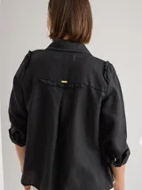 Camisa blanca de lino con cuello clásico, abotonada al frente con botones de madera, mangas largas con volados en los hombros y corte femenino.