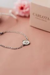 Pulsera de acero quirúrgico plateado con cadena de eslabones y dije circular blanco con borde negro y la letra "D" en negro.