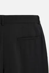 Pantalón negro de corte amplio, con pliegues frontales, bolsillos laterales y traseros de ojal. Incluye cinturón de la misma tela con hebilla metálica.