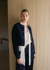 Chaqueta corta de jean azul oscuro, con cuello redondo, cierre frontal con botones y dos bolsillos de parche en la parte delantera.