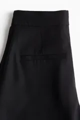 Pantalón culotte negro de tiro alto con cierre, botón y corchetes ocultos. Tiene bolsillos al bies y bolsillos traseros decorativos. Las piernas son amplias con pliegues adelante y atrás.