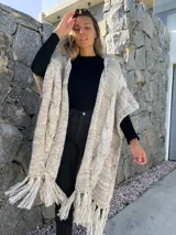 Ruana larga de lana merino color beige jaspeado, con diseño de trenzas y flecos en el borde inferior.