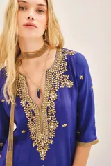 Vestido largo azul vibrante con escote en V profundo, mangas 3/4 y caída fluida. Presenta un elaborado bordado frontal en el escote con piedras, cintas e hilos de lurex en tonos dorados y plateados, con pequeños apliques florales dorados distribuidos en el cuerpo de la prenda. Incluye un accesorio colgante de cuentas doradas a modo de bufanda o detalle decorativo.