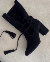 Botín de gamuza negro con taco cuadrado, caña media y tiras finas con borlas para atar alrededor del tobillo.