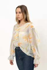 Blusa blanca traslúcida con estampado floral color naranja, cuello con volados y mangas largas abullonadas con puños elásticos.