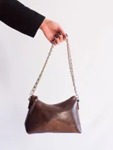 Cartera marrón de cuero con forma de media luna, forro interior y bolsillo con cierre. Tiene una correa de cadena metálica plateada.