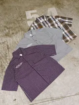 Camisa de manga corta con corte híbrido boxy fit/oversize, confeccionada en algodón. Disponible en tres diseños: a cuadros marrones, a rayas blancas y negras, y a rayas burdeos y blancas. Presenta bordados de diseño propio de la marca.