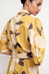 Vestido midi camisero color amarillo con estampado de flores blancas y hojas marrones. Tiene cuello solapa, mangas 3/4 con puños, cinturón de lazo ajustable y cierre frontal con botones.