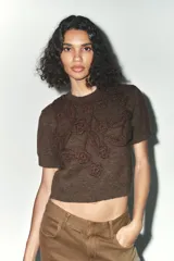 Sweater marrón de tejido de punto con cuello redondo y mangas cortas. Presenta apliques florales tejidos al crochet en el frente.