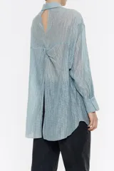Camisa larga de viscosa con textura, de corte recto, con cuello camisero, manga larga de puño con botón y cierre frontal con abotonadura. Presenta bolsillo de plastrón en el pecho y nudo decorativo en la parte trasera para dar volumen.