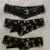 Cinto ancho de cuero negro con hebilla metálica plateada y detalles de tachas o argollas metálicas.