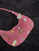 Cartera baguette de gamuza rosa con asa corta y múltiples apliques decorativos: botones, monedas, dijes de metal con forma de sol y caballo, y placa metálica con la marca "Agnes Lenoble".