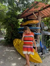 Vestido corto tejido con hilo de algodón a rayas horizontales en tonos celeste, azul, marrón, blanco, naranja y verde. Tiene cuello redondo y mangas cortas. El ruedo es desflecado.