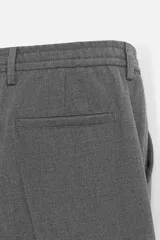 Pantalón gris de corte ancho con cintura elástica, pliegues frontales, bolsillos laterales y traseros de vivo. Incluye detalle de hebilla metálica en la trabilla y cierre frontal con cremallera y botón.