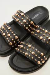 Sandalia tipo slide de color crudo, con dos tiras anchas adornadas con tachas doradas redondas de diferentes tamaños. La suela es gruesa y de goma.