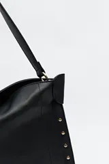 Bolso hobo color beige de símil cuero con textura suave y tachas metálicas en los bordes. Tiene correa al hombro y logo metálico en el frente.