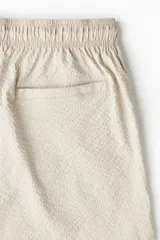 Short de baño color beige con textura de sirsaca, cintura elástica con cordón ajustable, bolsillos laterales y trasero ribeteado, aberturas laterales cortas y suspensor interior de malla.