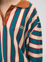 Chomba de corte oversize con cuello camisero, a rayas verticales en color marrón, lila y blanco. Tiene un logo bordado en el pecho y puños en color marrón.