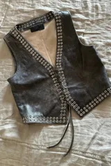 Chaleco negro de cuero con escote en V y cierre frontal con cordones. Presenta ojalillos metálicos plateados a lo largo de los bordes.