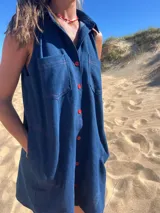 Túnica sin mangas de tela denim azul oscuro, con cuello camisero, cierre frontal de botones rojos y pespuntes en hilo rojo. Presenta bolsillos de parche en el pecho y bolsillos laterales.