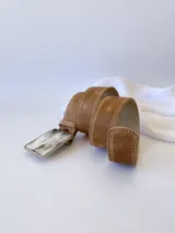 Cinto de cuero color marrón con forro de cuero y hebilla rectangular con diseño de guampa en blanco y negro con marco metálico plateado.