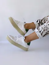 Zapatilla de cuero blanco con detalle verde en el talón y suela gruesa.