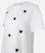 Remera baby look blanca de algodón con cuello redondo y mangas cortas. Presenta bordados de lacitos negros en el frente.