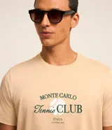 Remera color arena de algodón, con cuello redondo y mangas cortas. Estampa frontal con el texto "Monte Carlo Tennis Club" en color verde.